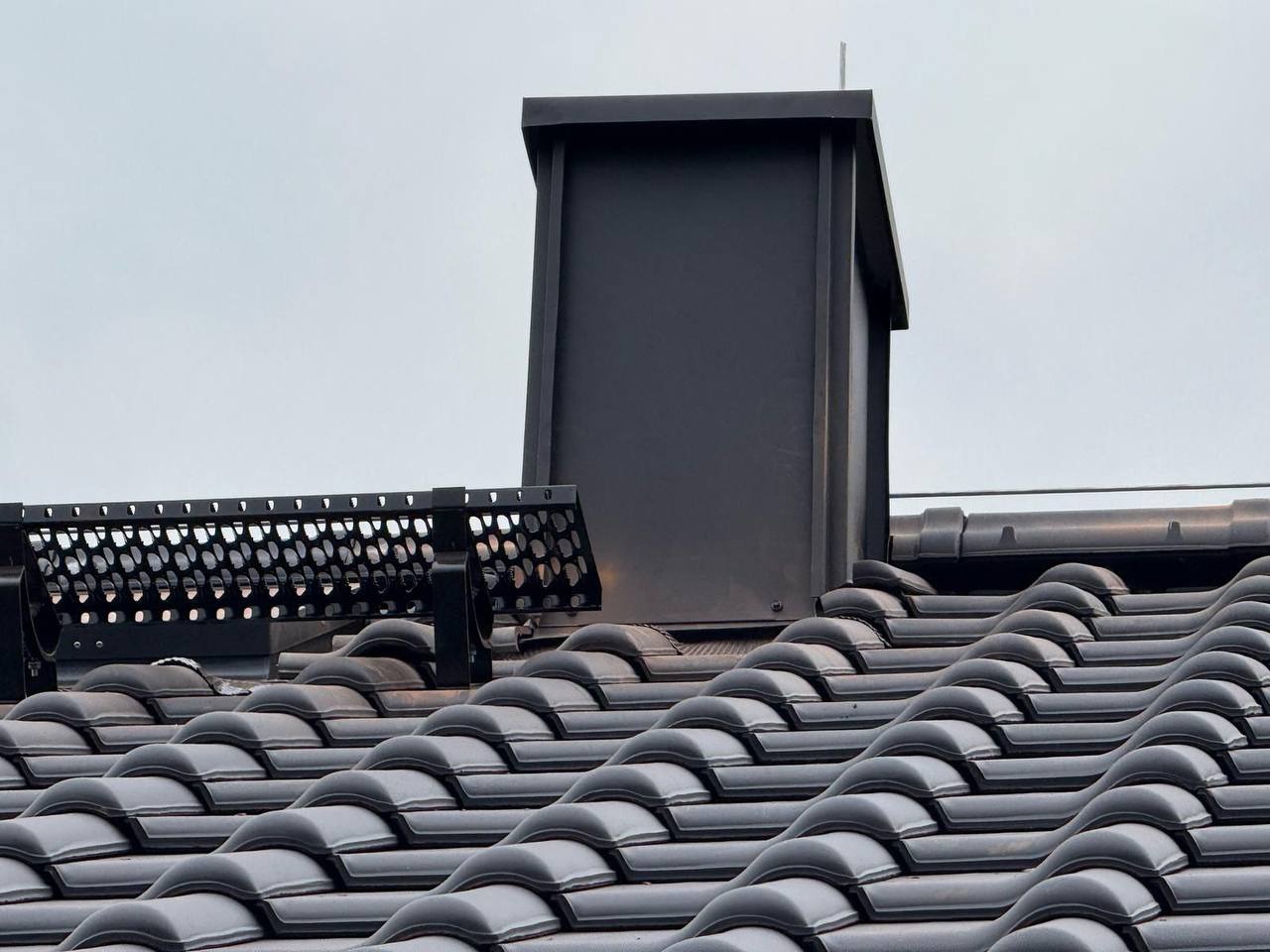 Czarny komin wentylacyjny z osłoną przeciwśnieżną na dachu pokrytym ciemną dachówką ceramiczną, widoczne z bliskiej perspektywy pod kątem.