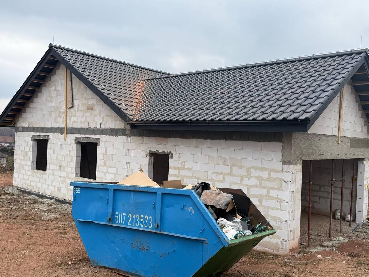Niedokończony dom z szarej cegły z ciemnoszarym dachem i niebieskim kontenerem na śmieci na pierwszym planie, w pochmurny dzień.