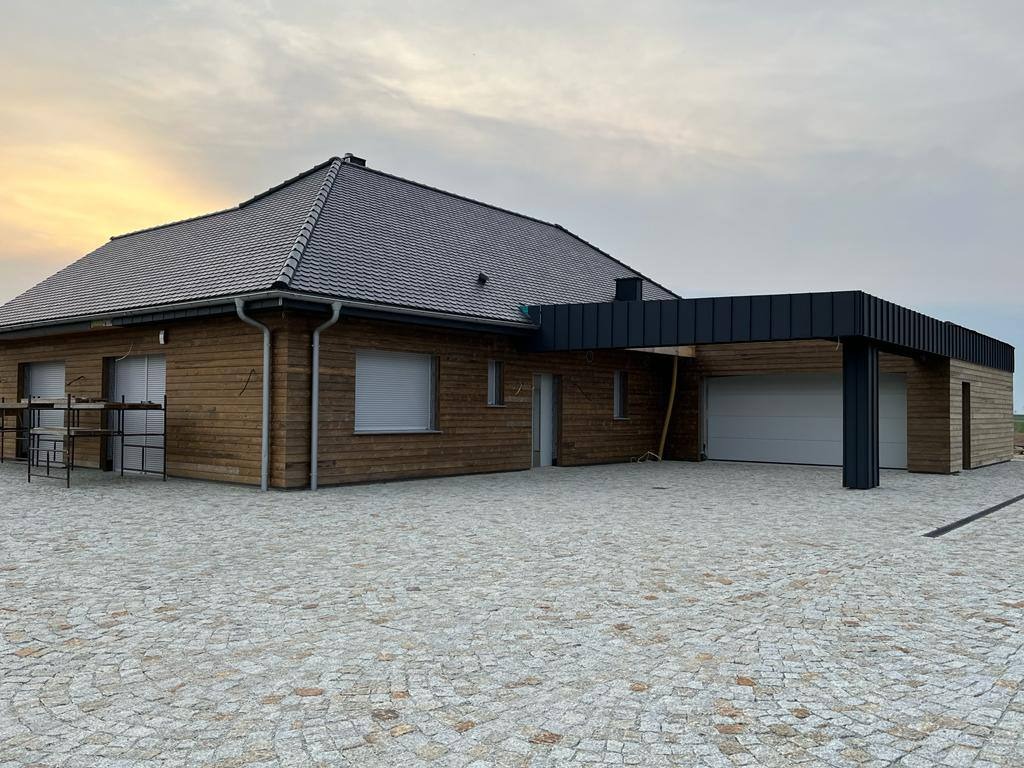 Nowoczesny dom z drewnianą elewacją i garażem, wykończony kostką brukową. Widoczne okna z roletami i ciemna dachówka. Ujęcie z poziomu gruntu, pochmurne niebo w tle.