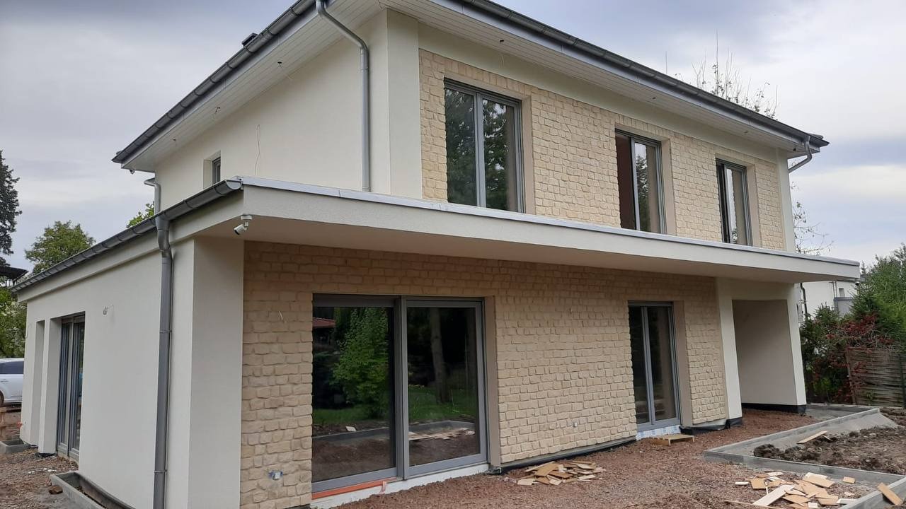 Dwukondygnacyjny dom z jasną elewacją, fragmentami wykończony cegłą. Widoczne okna z szarymi ramami i elementy orynnowania. Teren wokół domu częściowo zagospodarowany.