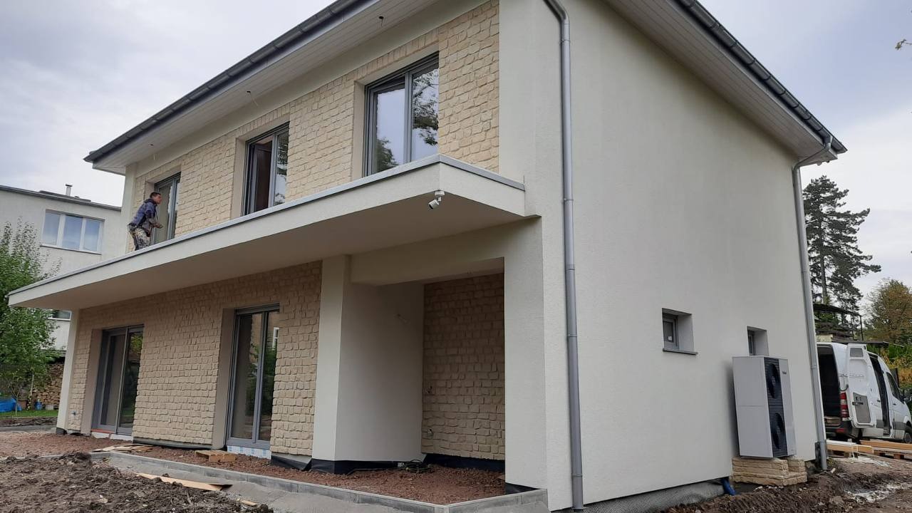 Dwukondygnacyjny dom z elewacją z jasnej cegły i białym tynkiem. Mężczyzna na dachu czyści okno. Widoczna pompa ciepła i częściowo samochód dostawczy.