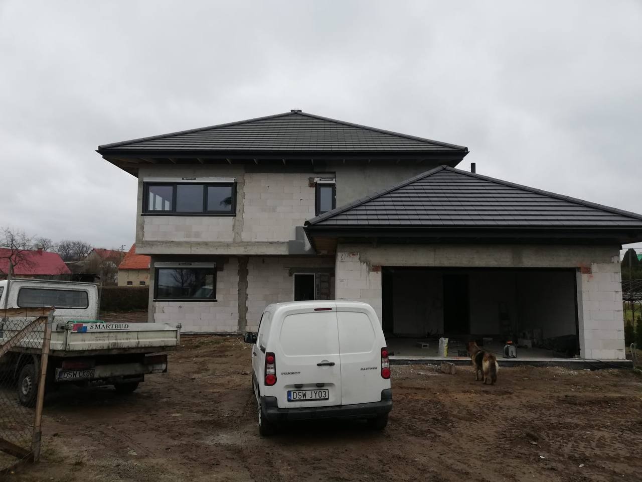 Surowy dom z szarej cegły z ciemnym dachem, widoczne okna i brama garażowa, na pierwszym planie samochody dostawcze i pies. Etap budowy.