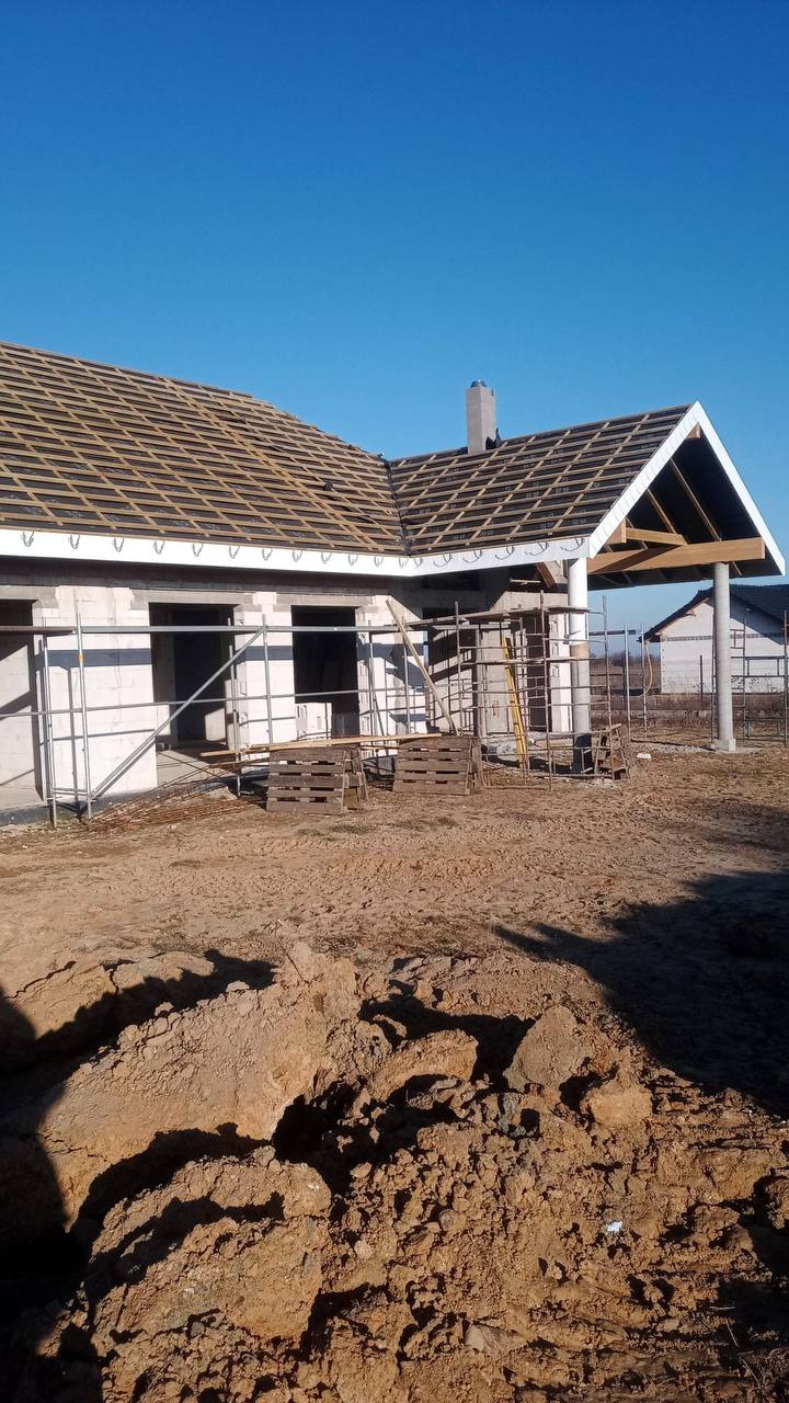 Budowa domu jednorodzinnego w stanie surowym otwartym. Widoczne ściany z bloczków, więźba dachowa z łatami i komin. Na pierwszym planie hałda ziemi.