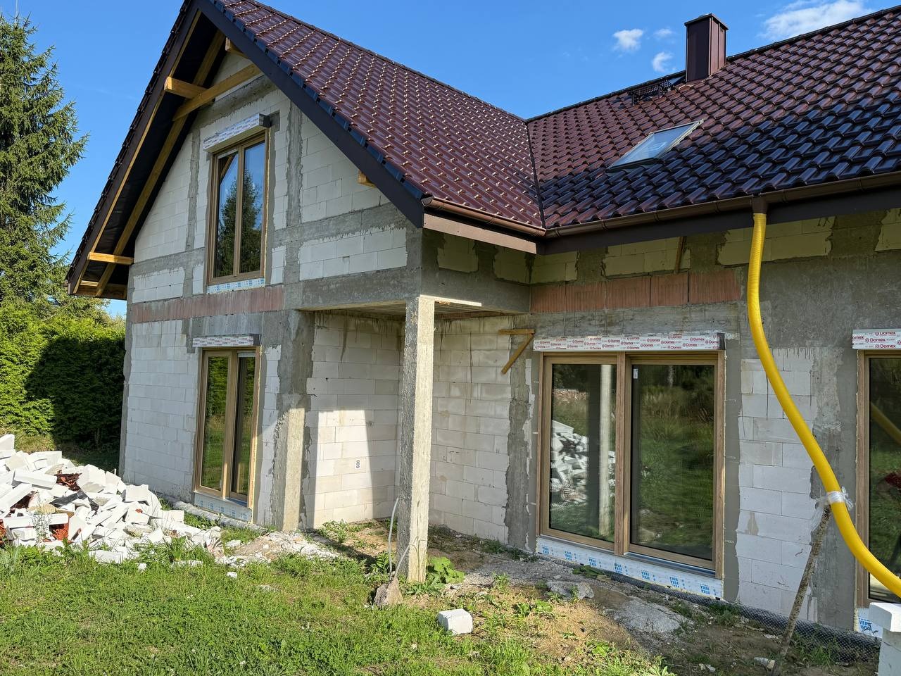 Niedokończony dom z brązowym dachem i oknami w drewnianych ramach. Na trawie leżą białe bloczki. Żółty wąż przy ścianie. Widoczne elementy konstrukcyjne.