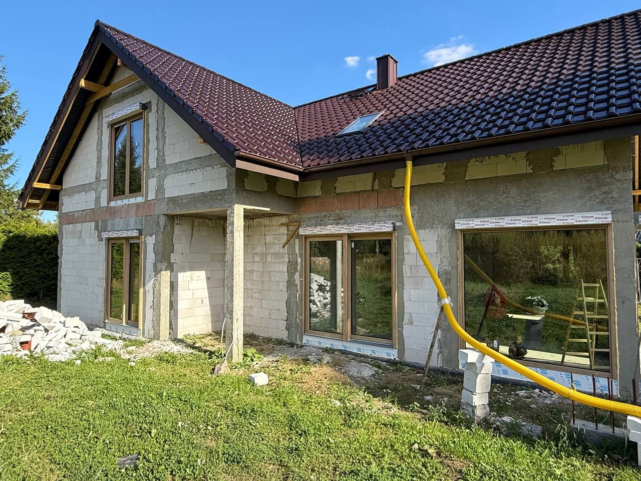 Surowy stan domu z pustaków, z brązowym dachem i oknami w drewnianych ramach. Żółty przewód wentylacyjny biegnie wzdłuż ściany. Widoczne materiały budowlane.