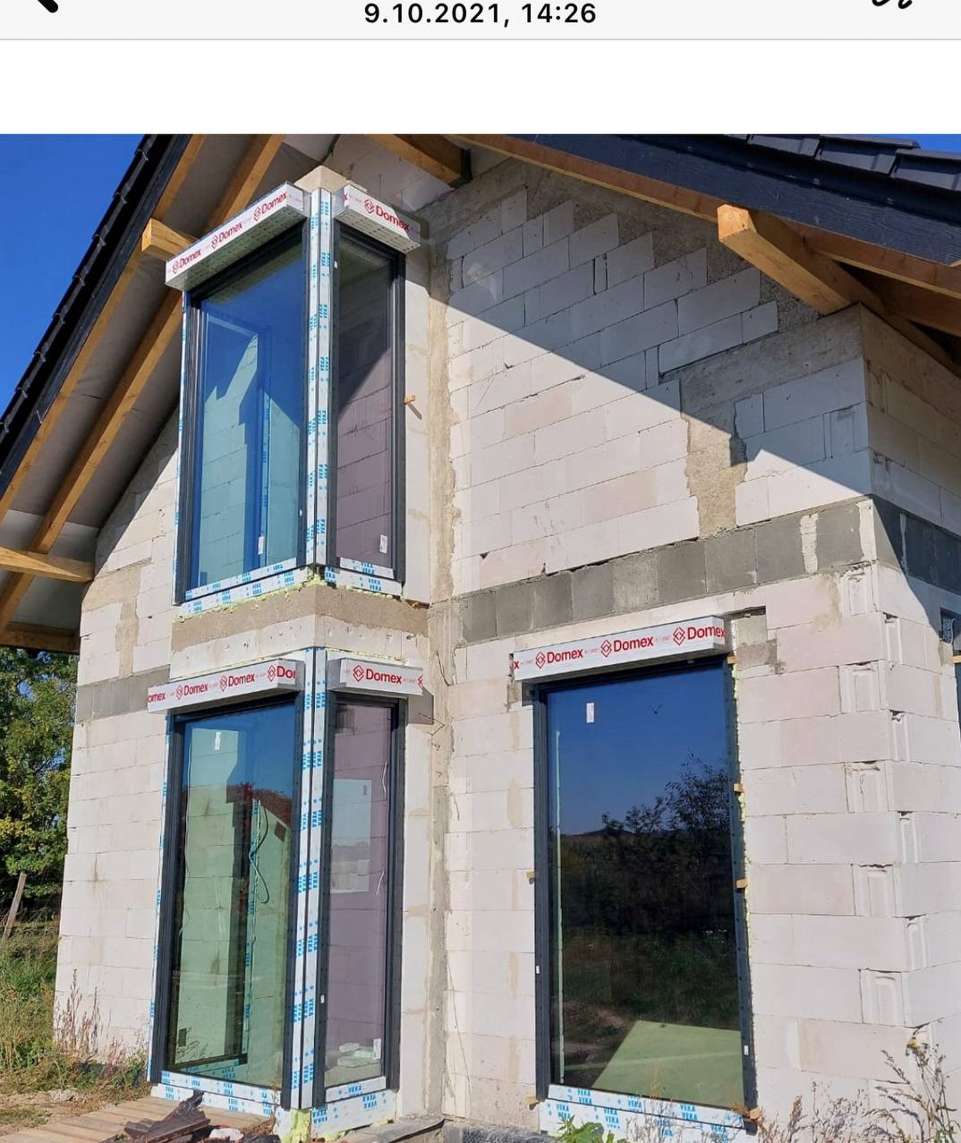 Surowy stan domu z białego bloczka z zamontowanymi oknami z czarnymi ramami, zabezpieczonymi folią. Widoczne elementy dachu i drewniane belki.