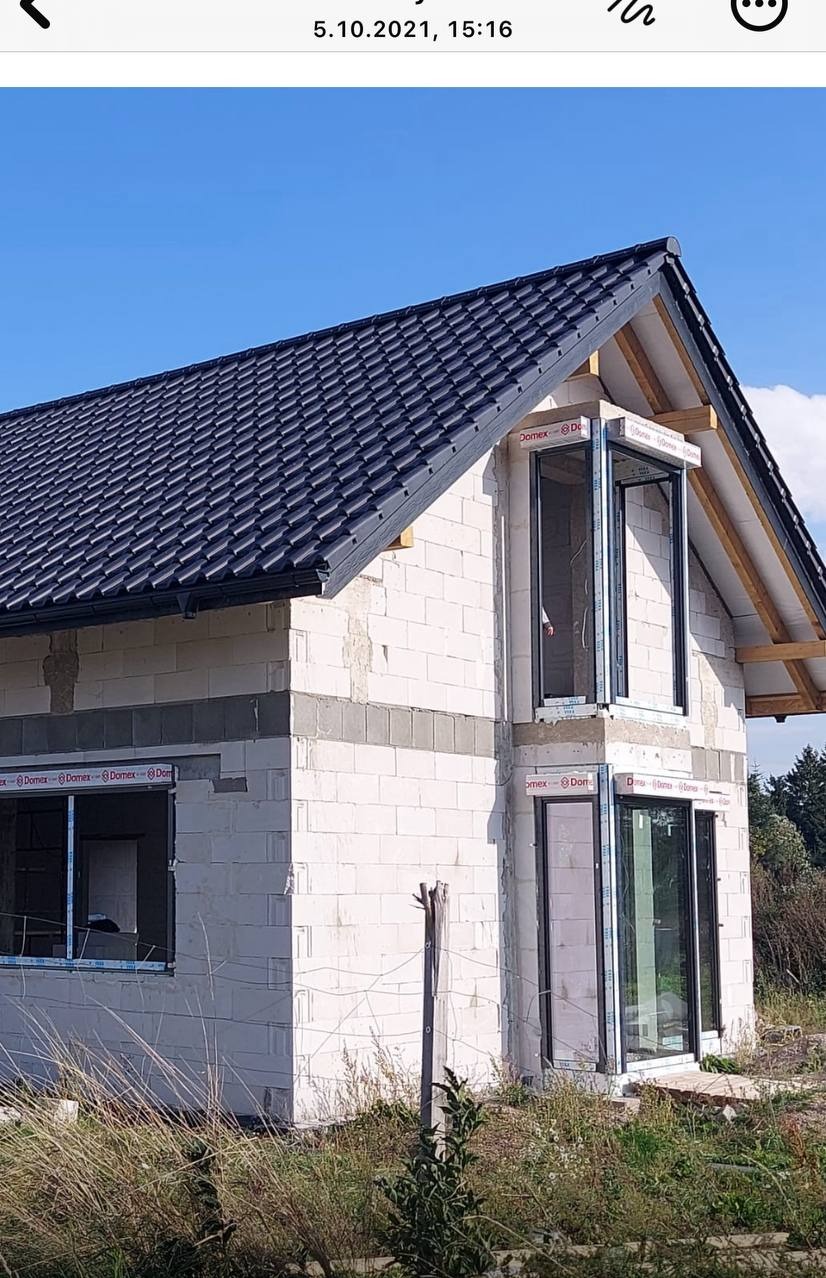 Surowy stan domu z białego bloczka, widoczne okna z czarnymi ramami i ciemny dach. Budynek w trakcie budowy, na tle błękitnego nieba i zieleni.