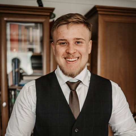 Uśmiechnięty mężczyzna w eleganckiej kamizelce i krawacie, z brodą, na tle regału z książkami. Portret.