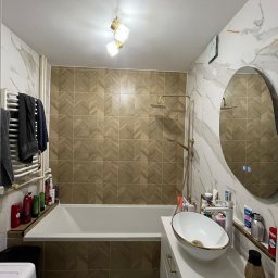 Oskar - Nowoczesna łazienka z beżowo-białymi płytkami, owalnym lustrem z podświetleniem LED, białą umywalką na szafce i złotą armaturą. Widoczny grzejnik drabinkowy i wanna.