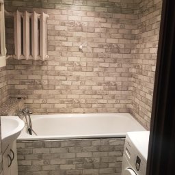 Oskar - Wykończona łazienka z wanną, umywalką i pralką. Ściany wyłożone tapetą imitującą cegłę, grzejnik żeberkowy. Mozaika na podłodze. Widok zza framugi drzwi.