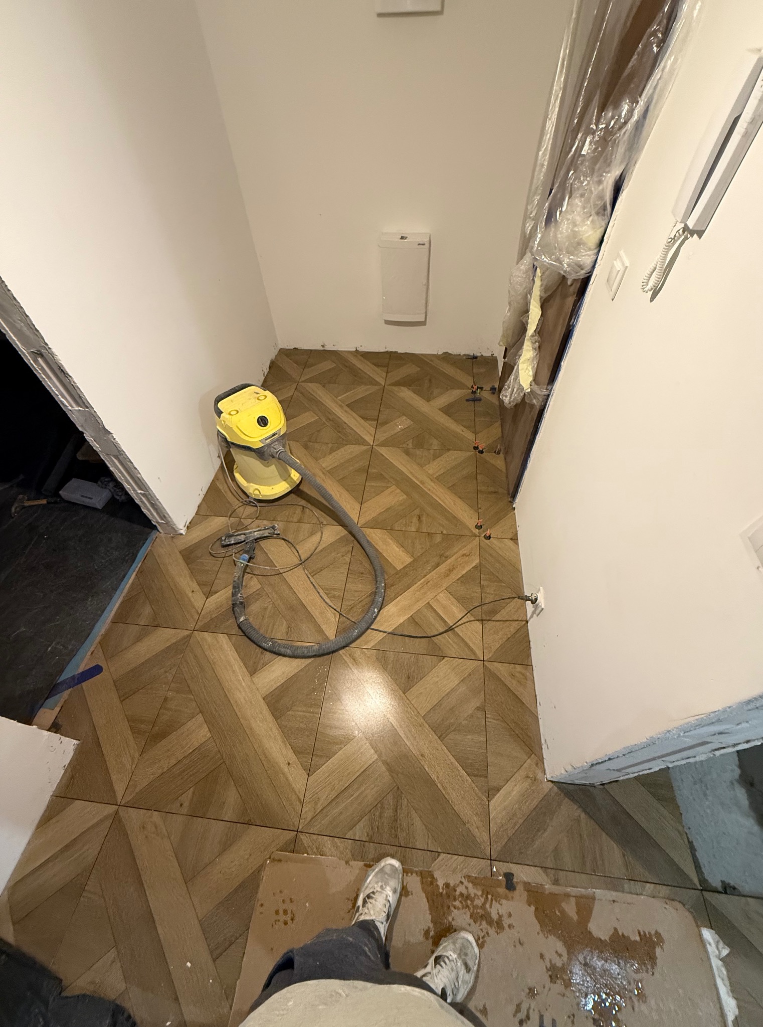 Układanie parkietu ceramicznego w korytarzu, widoczny odkurzacz przemysłowy i fragment osoby. Wzór jodełki, ciepłe odcienie brązu. Etap prac wykończeniowych.