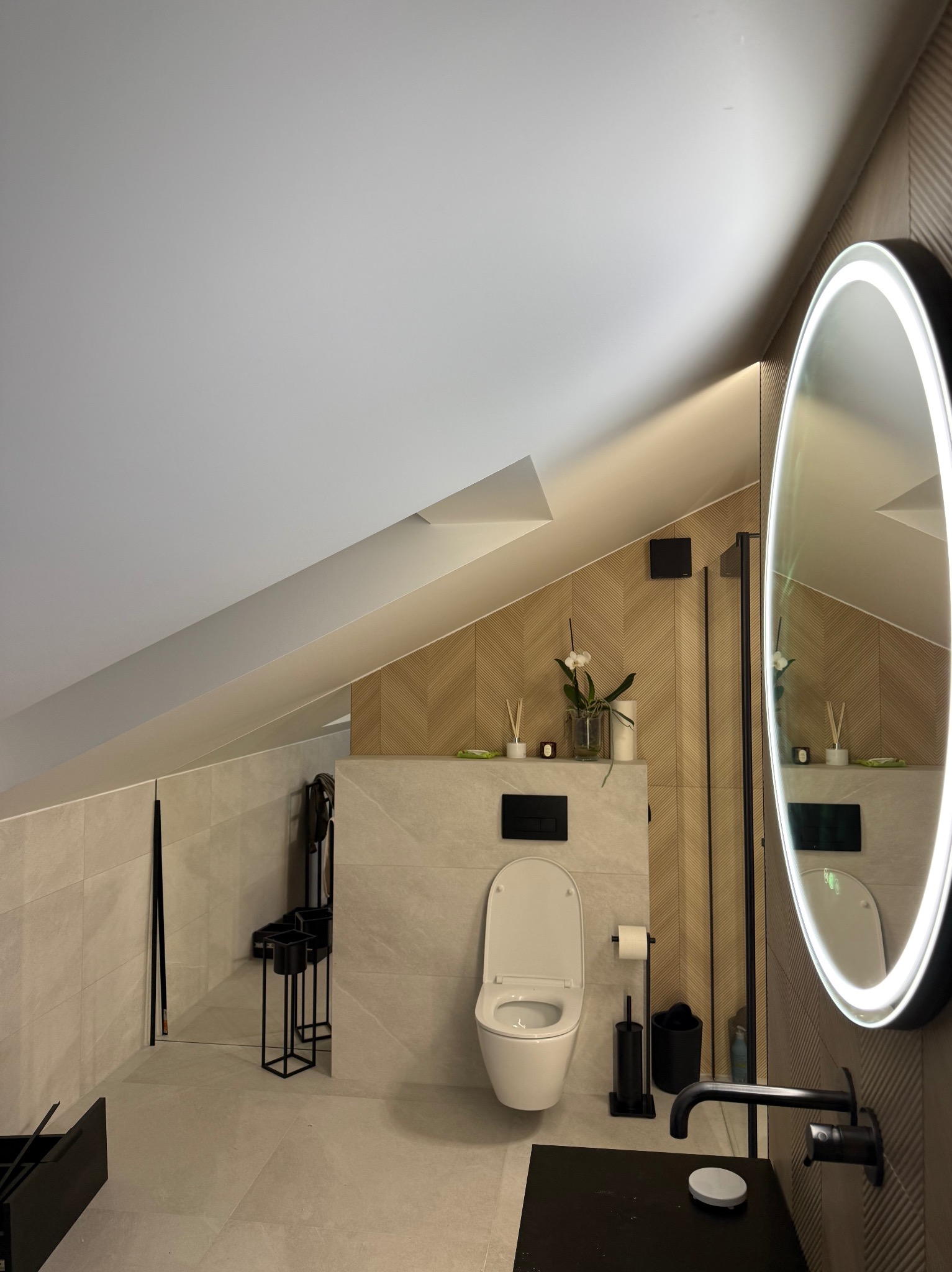 Nowoczesna łazienka na poddaszu z beżowymi płytkami, czarną armaturą i okrągłym lustrem z podświetleniem LED. Uwagę przykuwa skośny sufit i drewniane panele w jodełkę.