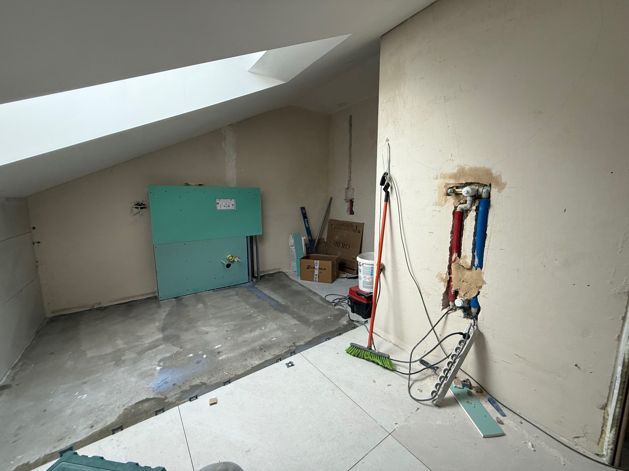 Poddasze w trakcie prac wykończeniowych: płyty GK na ścianie, częściowo ułożone płytki podłogowe, instalacja wodna i elektryczna w ścianie. Widoczne narzędzia i materiały budowlane.