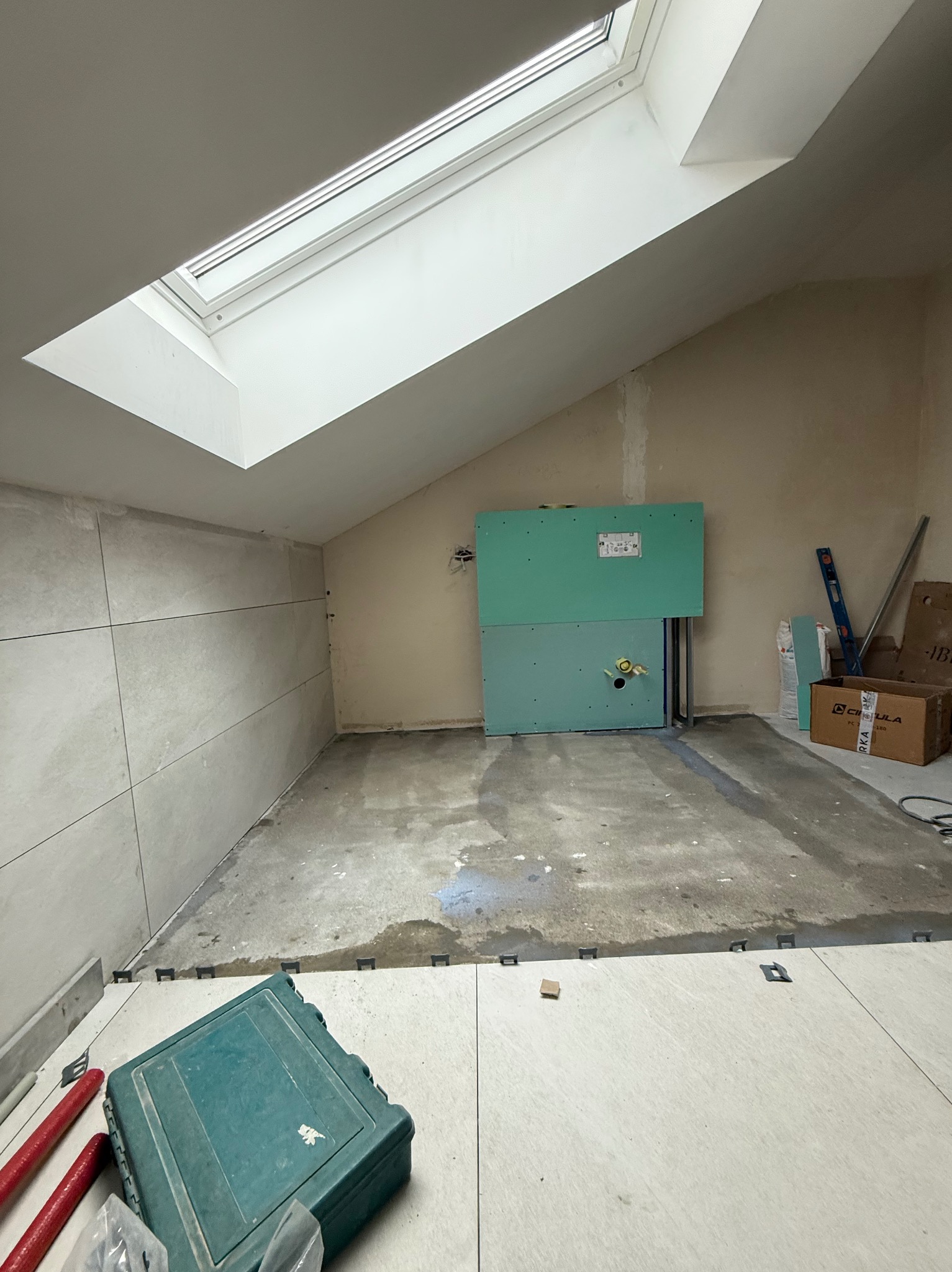 Poddasze w trakcie prac. Częściowo ułożone płytki na ścianie i podłodze, widoczny stelaż podtynkowy WC z płyt karton-gips, narzędzia i okno dachowe.