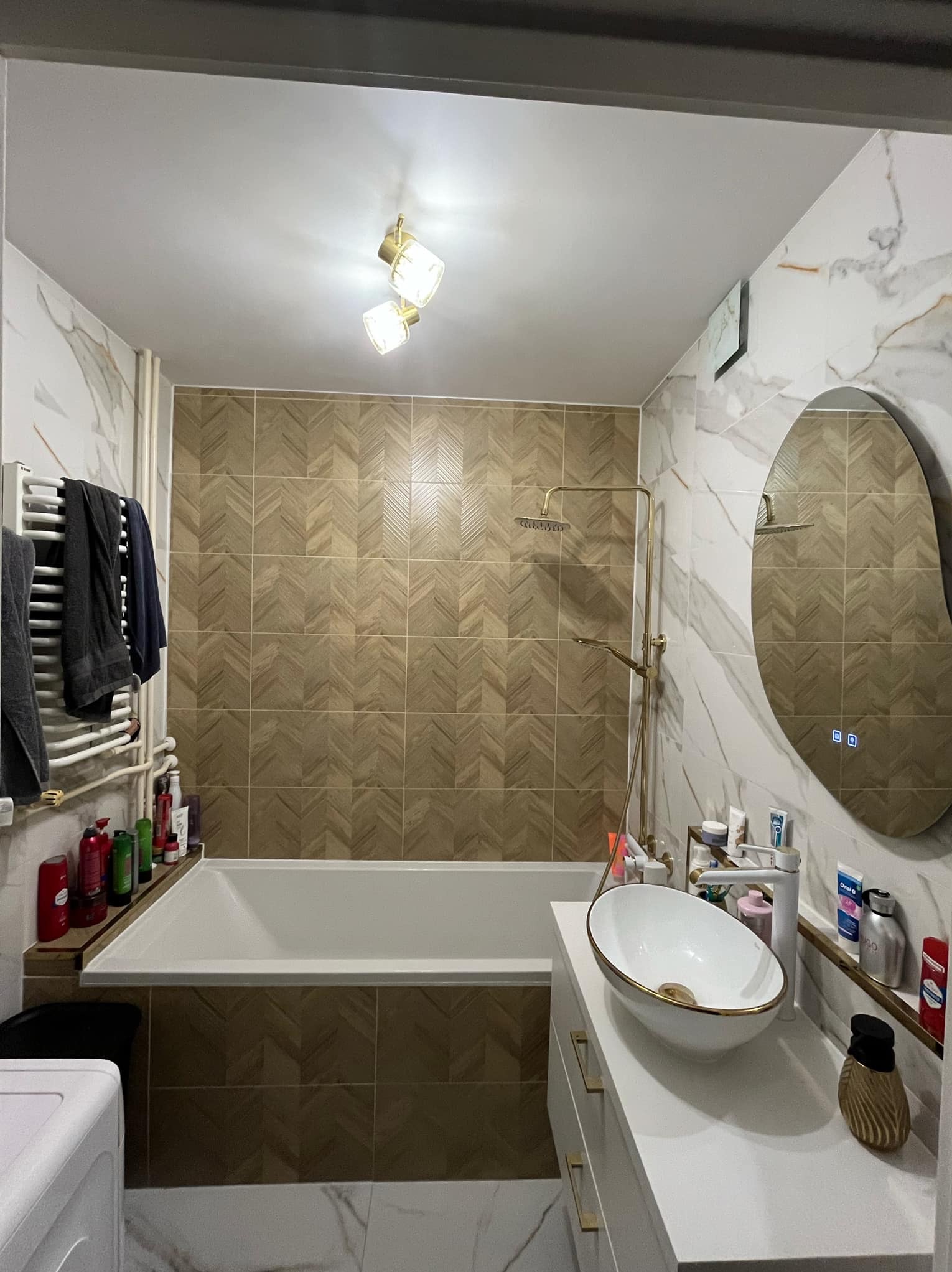 Nowoczesna łazienka z beżowo-białymi płytkami, owalnym lustrem z podświetleniem LED, białą umywalką na szafce i złotą armaturą. Widoczny grzejnik drabinkowy i wanna.