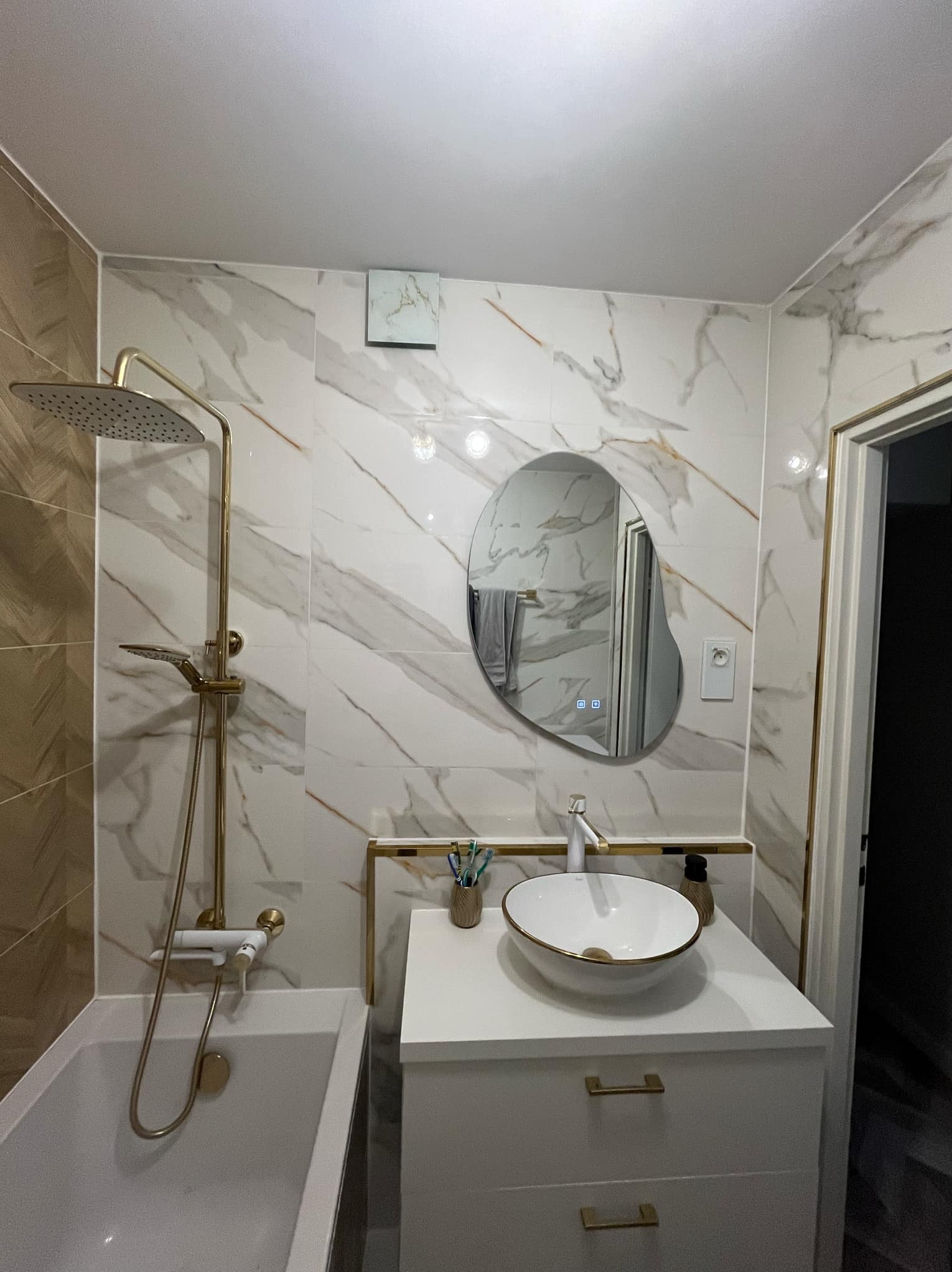 Elegancka łazienka z marmurowymi płytkami, złotą armaturą i asymetrycznym lustrem z podświetleniem LED. Nowoczesny design z dbałością o detale.