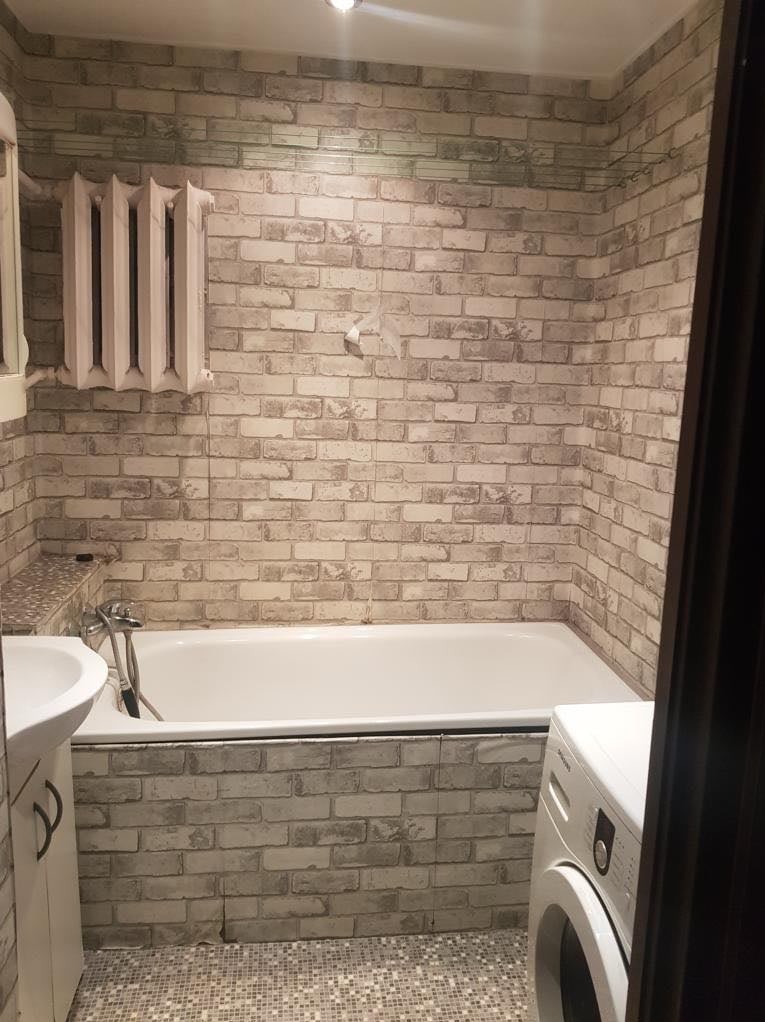 Wykończona łazienka z wanną, umywalką i pralką. Ściany wyłożone tapetą imitującą cegłę, grzejnik żeberkowy. Mozaika na podłodze. Widok zza framugi drzwi.