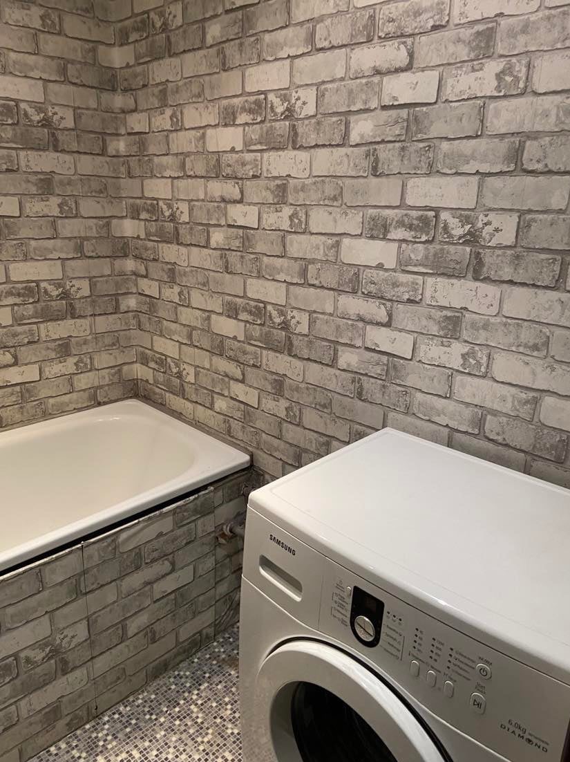 Łazienka z wanną i pralką Samsung, ściany wyłożone tapetą imitującą szarą cegłę. Podłoga wyłożona mozaiką. Ujęcie z niskiego kąta.