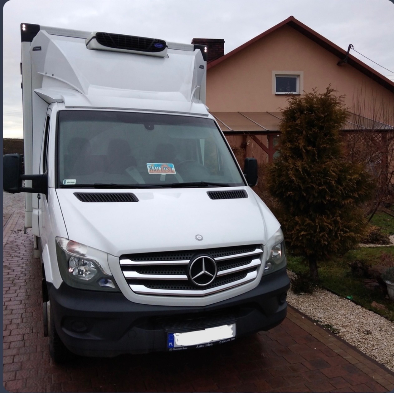 Biały Mercedes Sprinter z chłodnią na tle domu jednorodzinnego, zaparkowany na brukowanej nawierzchni. Widoczny przód pojazdu z logo Mercedes-Benz.