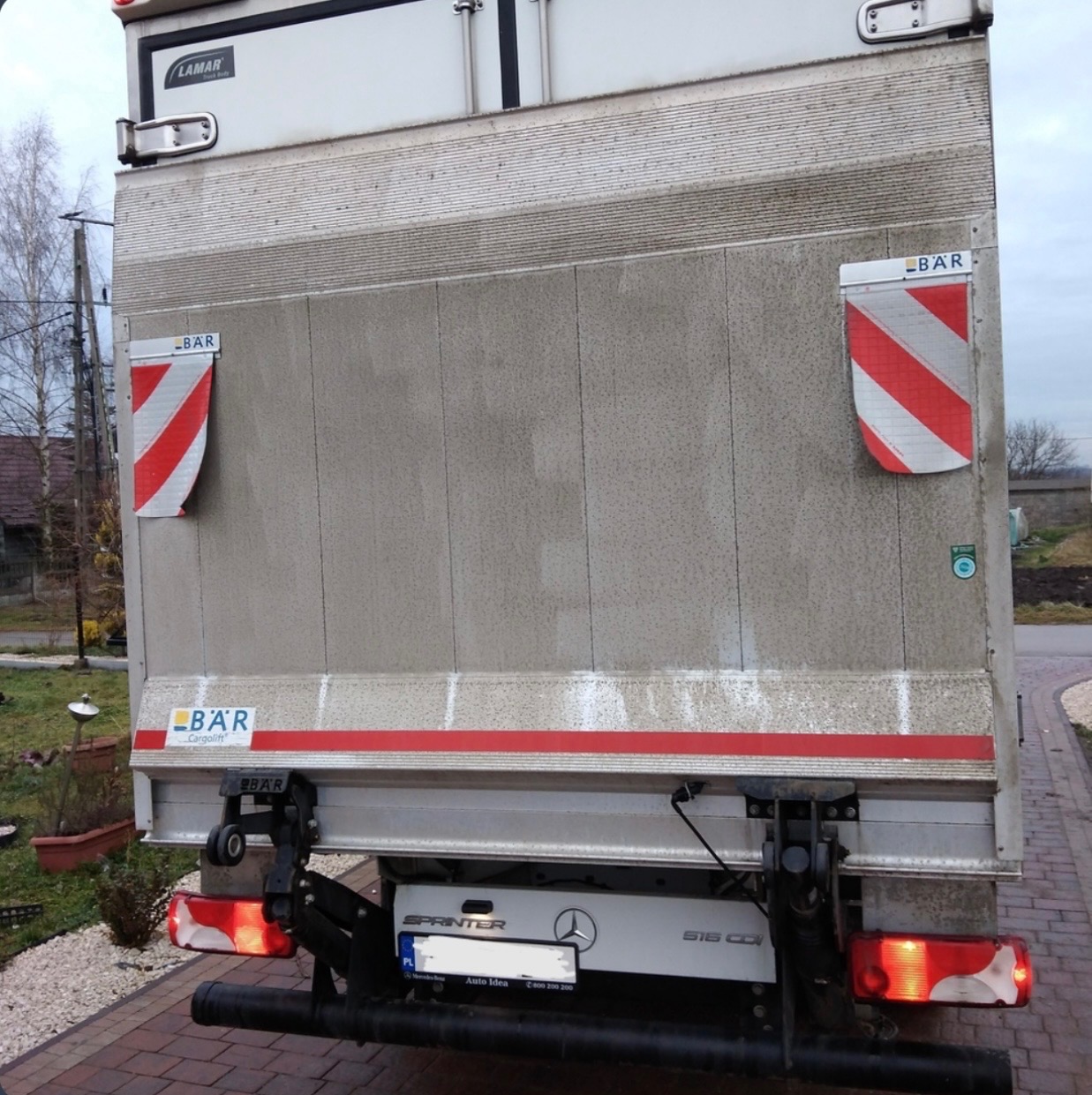 Tył brudnego busa transportowego Mercedes Sprinter z windą załadowczą BAR, widoczne światła, tablica rejestracyjna i elementy konstrukcyjne.