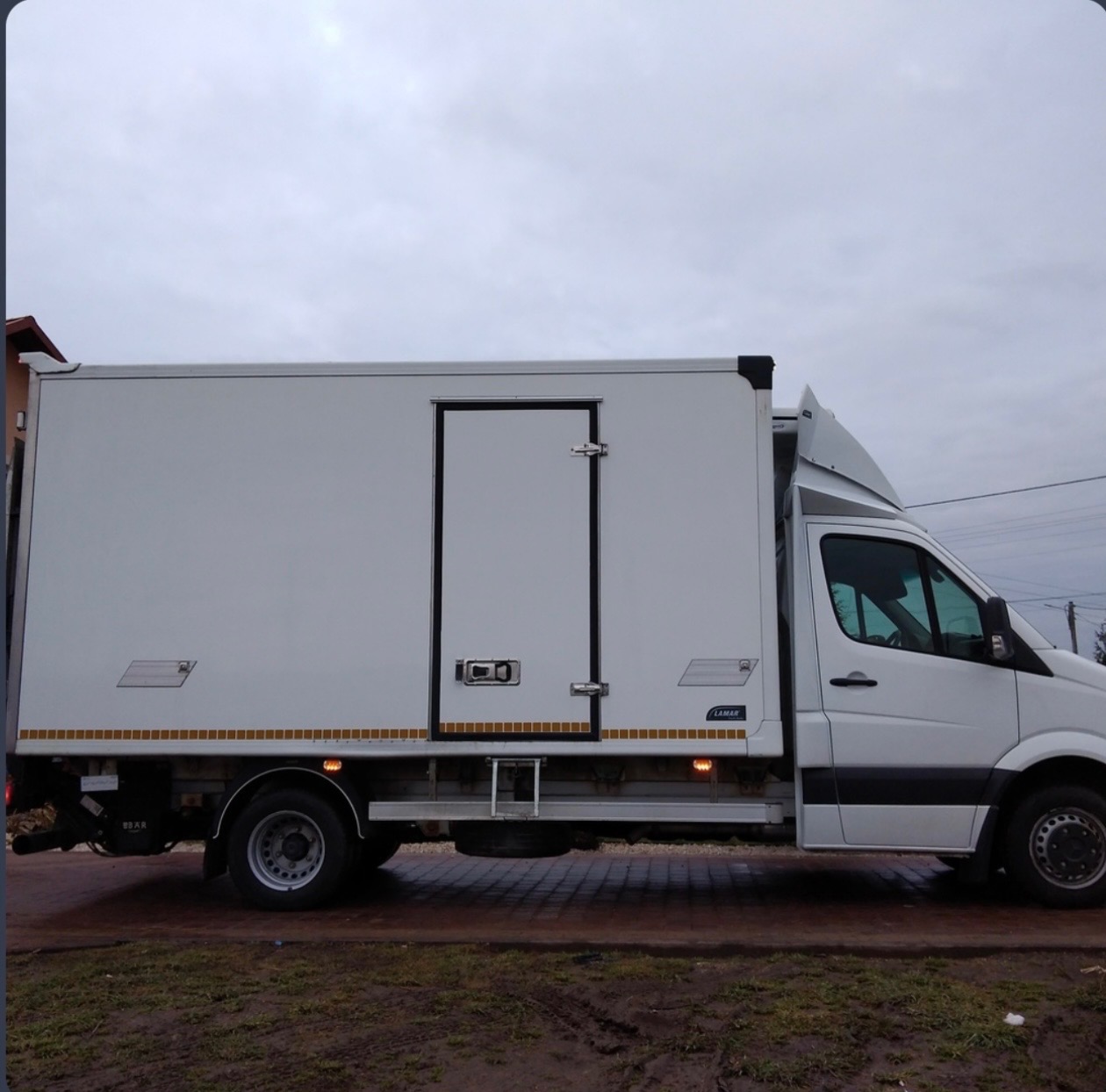 Biały samochód dostawczy typu furgon z zabudową kontenerową, zaparkowany na kostce brukowej, widok z boku, pochmurne niebo w tle.