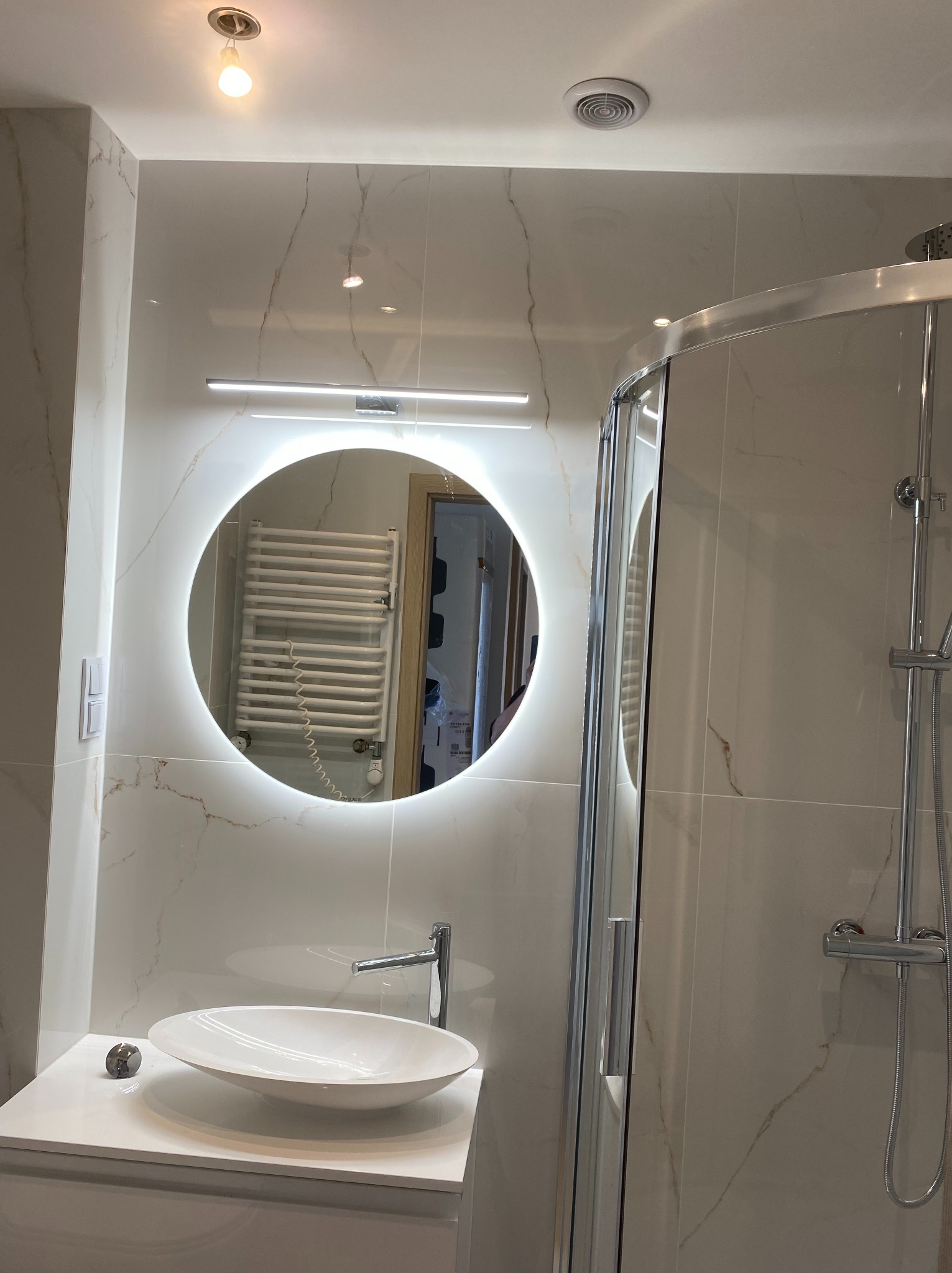 Nowoczesna łazienka z okrągłym lustrem LED, białą umywalką na szafce i kabiną prysznicową. Marmurowe płytki na ścianach. Widoczny grzejnik w odbiciu lustra.