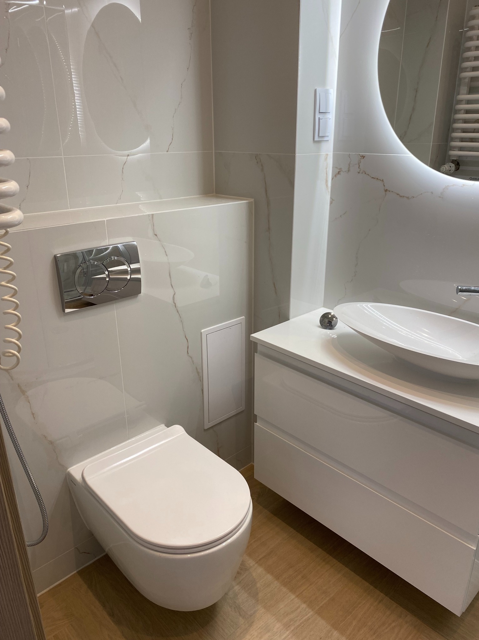 Nowoczesna łazienka z podwieszaną toaletą, umywalką na szafce i okrągłym lustrem z podświetleniem LED. Białe płytki imitujące marmur na ścianach i drewnopodobna podłoga.