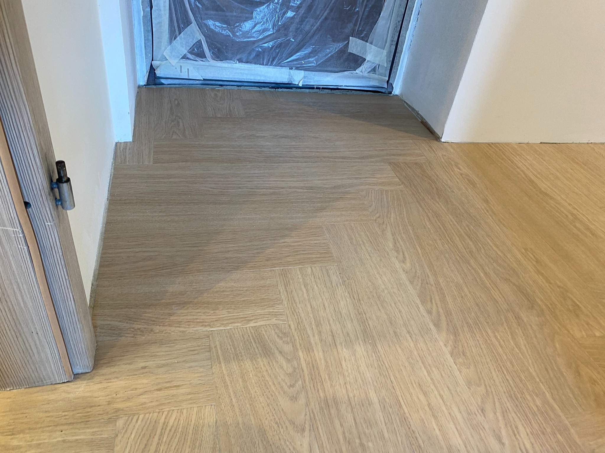 Ułożone panele podłogowe w jodełkę, widok z góry, jasne drewno, fragment drzwi po lewej, zabezpieczone folią drzwi balkonowe w tle.