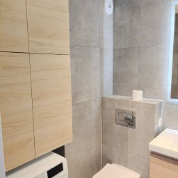 Dimfix - Nowoczesna łazienka z szarymi płytkami, podwieszaną toaletą i pralką pod zabudową z drewnianymi szafkami. Minimalistyczny design z akcentem na funkcjonalność i estetykę.