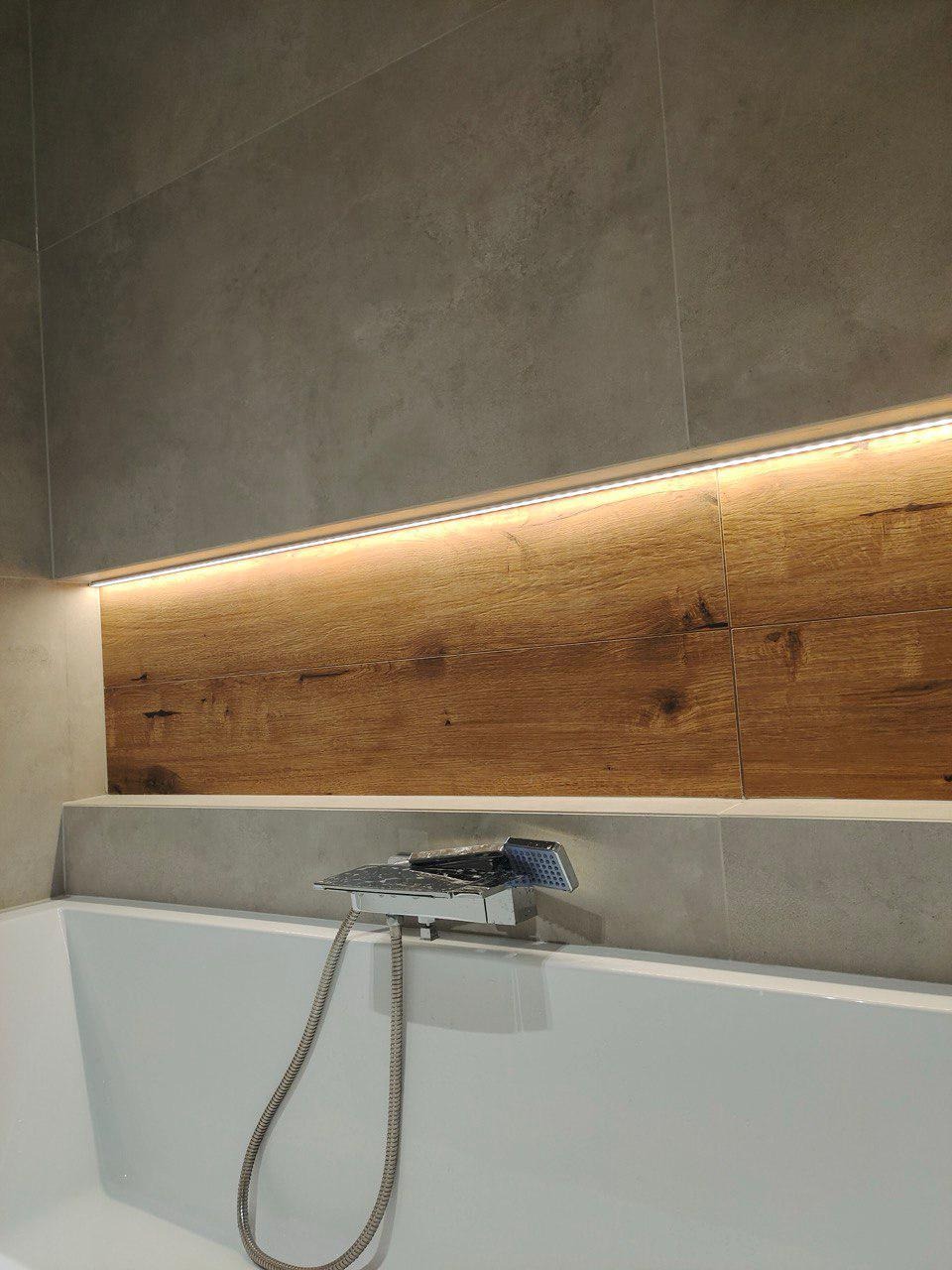 Nowoczesna łazienka: biała wanna z chromowaną baterią, drewniana wnęka z oświetleniem LED i szare płytki. Minimalistyczny design, ciepłe światło.