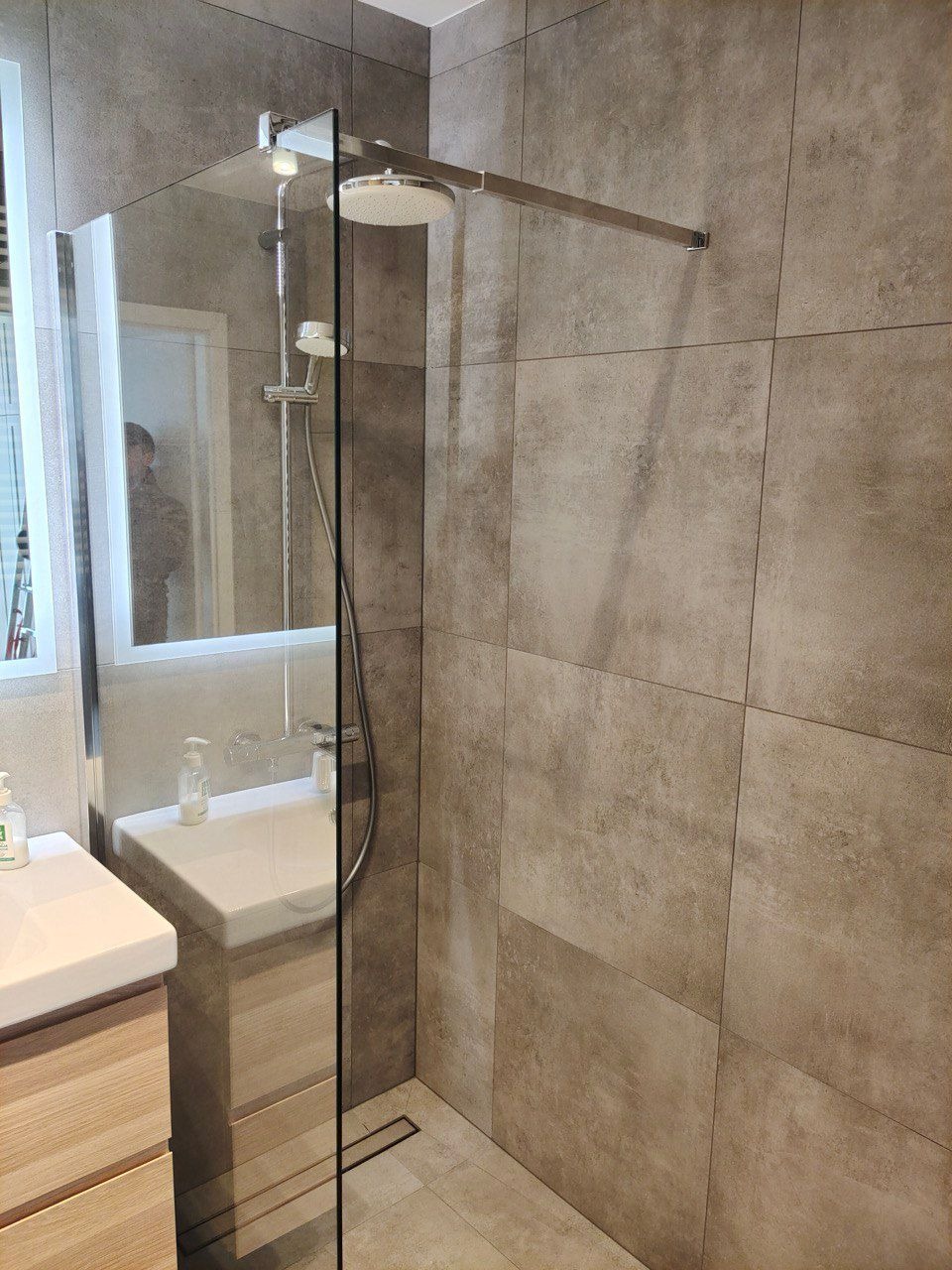 Nowoczesna kabina prysznicowa walk-in z deszczownicą i szklaną ścianką, wykończona płytkami w odcieniach szarości. Minimalistyczna umywalka z szafką w tle.