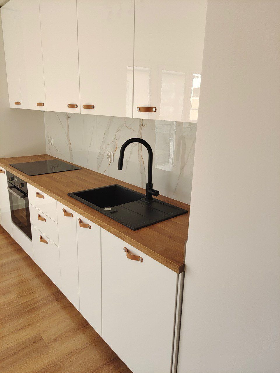 Biała kuchnia z drewnianym blatem, czarnym zlewem i baterią. Górne szafki lakierowane na wysoki połysk, marmurowy backsplash. Minimalistyczny design z akcentami w postaci skórzanych uchwytów.