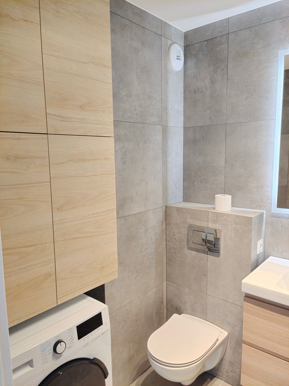 Nowoczesna łazienka z szarymi płytkami, podwieszaną toaletą i pralką pod zabudową z drewnianymi szafkami. Minimalistyczny design z akcentem na funkcjonalność i estetykę.