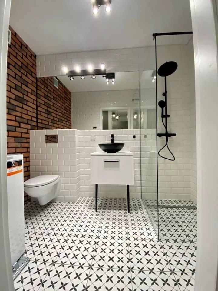 Nowoczesna łazienka z ceglaną ścianą, białą armaturą i czarnymi akcentami. Uwagę przykuwają wzorzyste płytki podłogowe i minimalistyczna kabina prysznicowa.