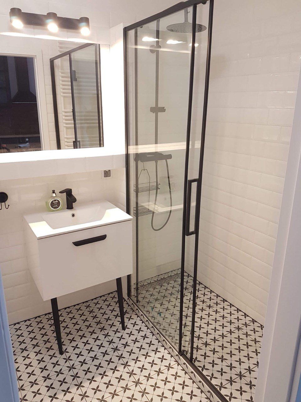 Nowoczesna łazienka z kabiną prysznicową typu walk-in z czarnymi profilami, białą umywalką na czarnych nóżkach i wzorzystą podłogą w czarno-białe krzyżyki.