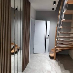 Damian Kowalczyk - Nowoczesny przedpokój z drewnianą ścianką działową z lustrami, schodami z drewnianymi stopniami i białymi drzwiami z czarnymi akcentami. Szara podłoga z płytek.