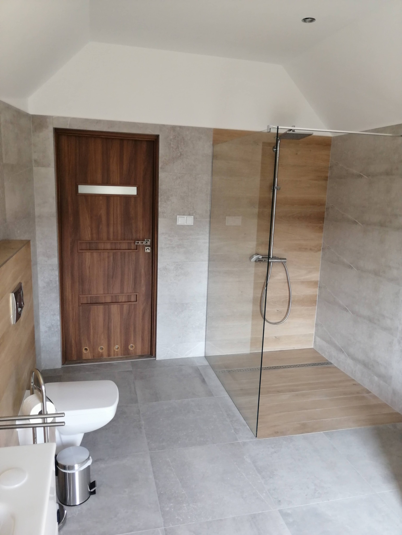 Nowoczesna łazienka po remoncie w Szczecinie: szare płytki, kabina walk-in z drewnopodobną podłogą, drewniane drzwi z przeszkleniem, minimalistyczny design.