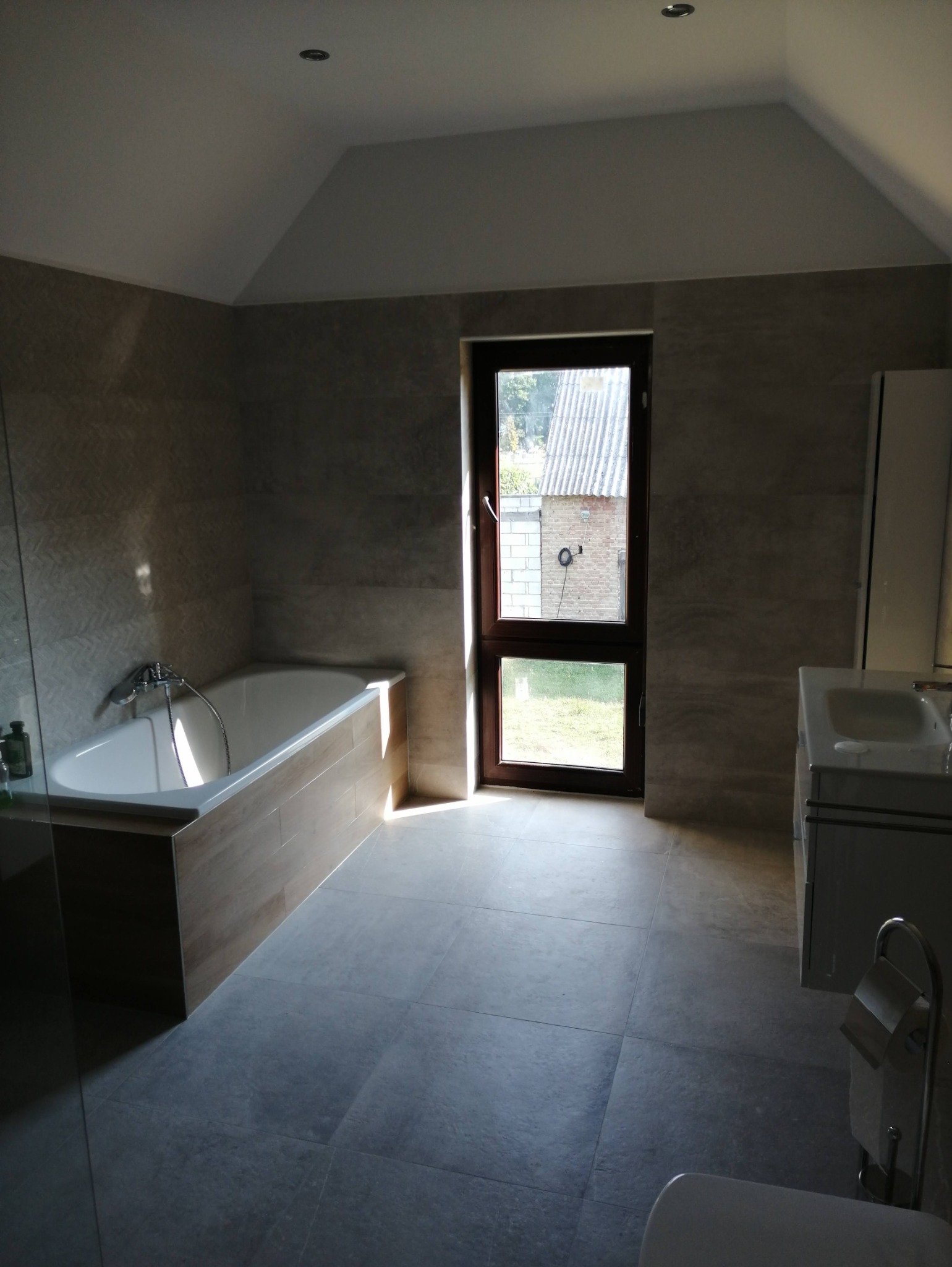 Jasna łazienka na poddaszu z oknem, wykończona płytkami w odcieniach szarości i beżu. Widoczna wanna, umywalka z szafką i fragment toalety.