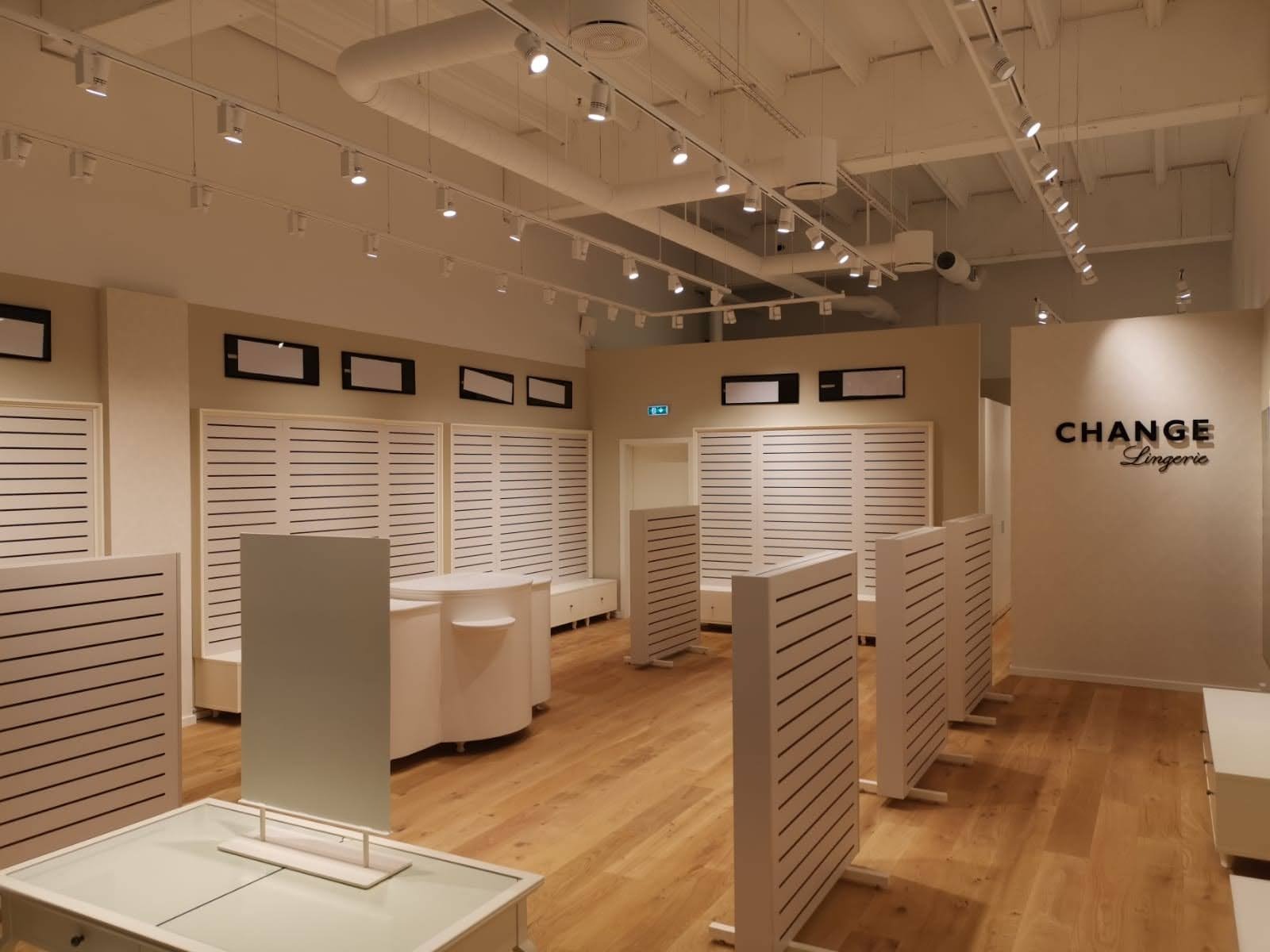 Jasne wnętrze sklepu odzieżowego z drewnianą podłogą, białymi meblami i oświetleniem punktowym. Widoczne regały z poziomymi listwami i logo marki na ścianie.