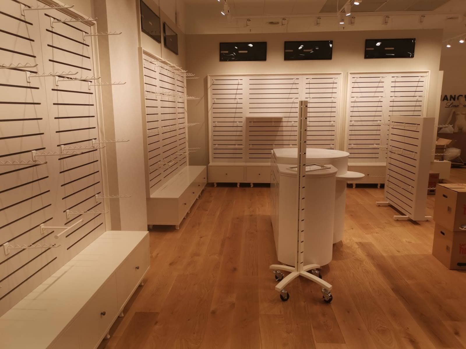 Jasne wnętrze sklepu odzieżowego z drewnianą podłogą, białymi panelami ściennymi z wieszakami i mobilnym, okrągłym stołem ekspozycyjnym na kółkach.