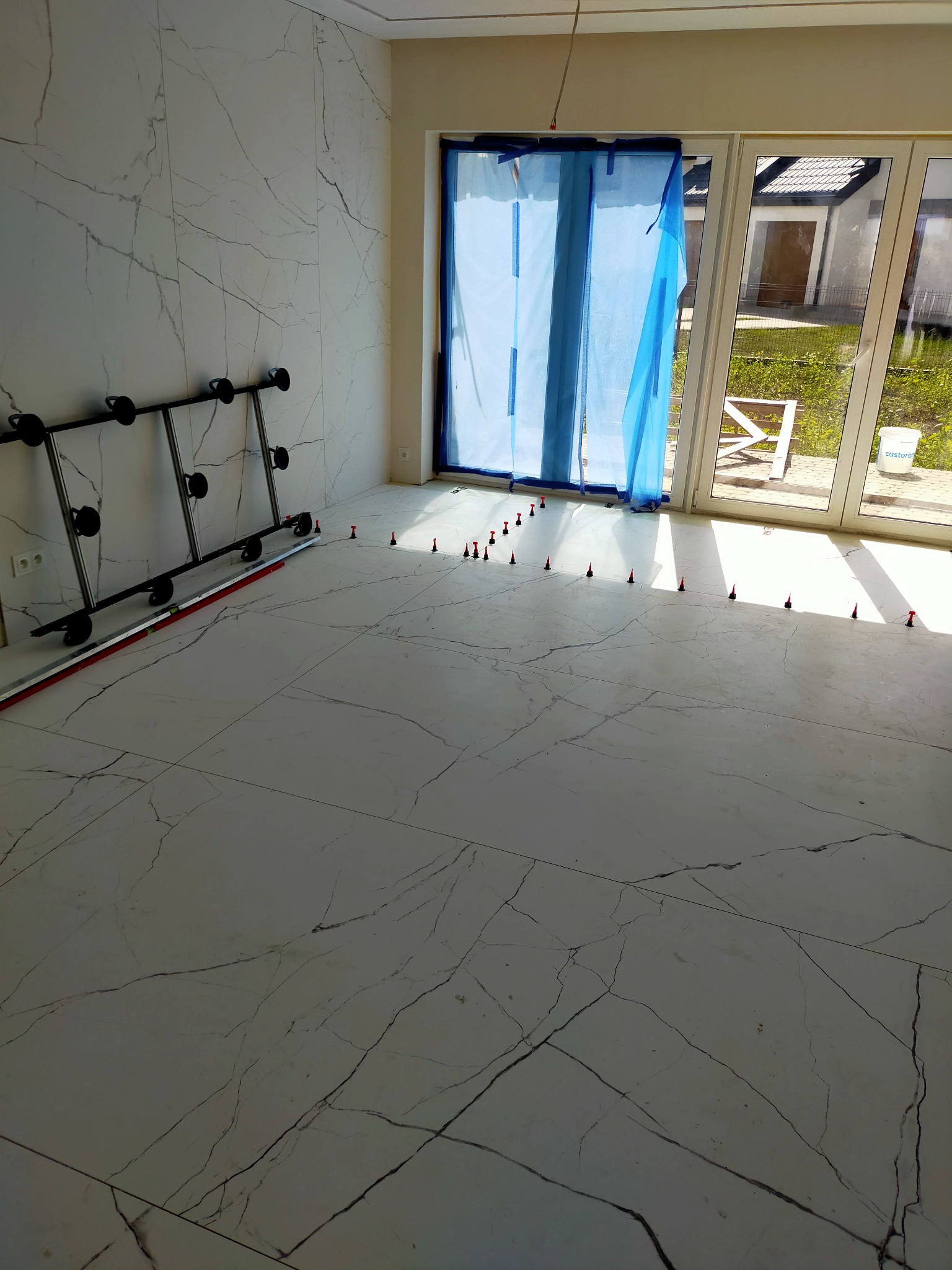 Układanie płytek imitujących marmur na podłodze i ścianie. Widoczne przyssawki do przenoszenia płytek i system poziomowania płytek. Okno zabezpieczone folią.