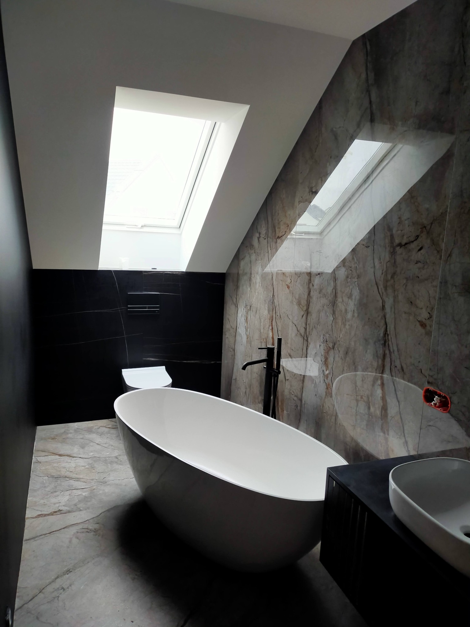 Nowoczesna łazienka na poddaszu z oknami dachowymi, marmurowymi płytkami i wolnostojącą wanną. Minimalistyczny design z czarnymi akcentami i elegancką armaturą.