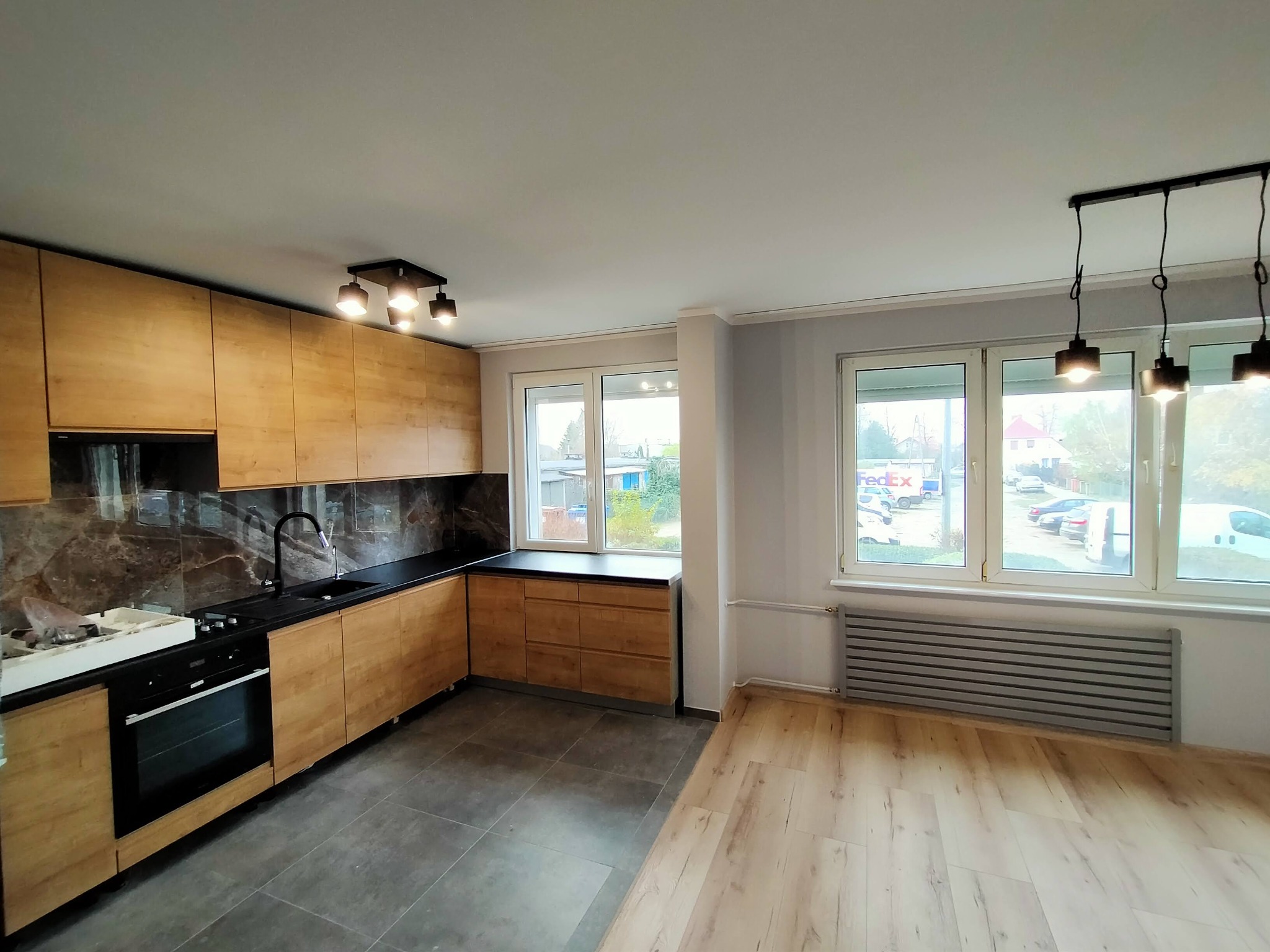 Jasna kuchnia z drewnianymi szafkami i czarnym blatem, widok na okno z grzejnikiem, podłoga z płytek i paneli. Nowoczesne oświetlenie sufitowe.