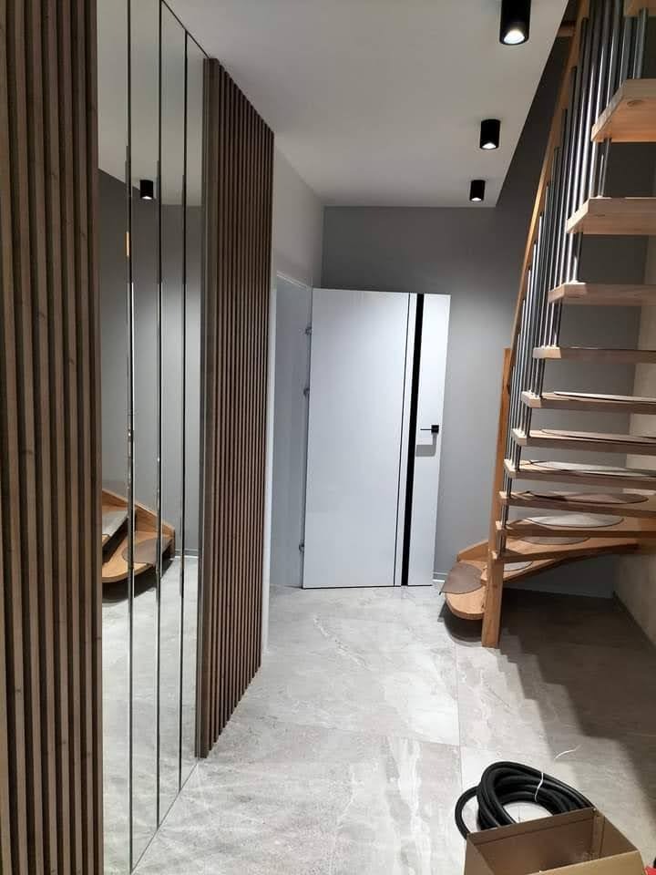 Nowoczesny przedpokój z drewnianą ścianką działową z lustrami, schodami z drewnianymi stopniami i białymi drzwiami z czarnymi akcentami. Szara podłoga z płytek.