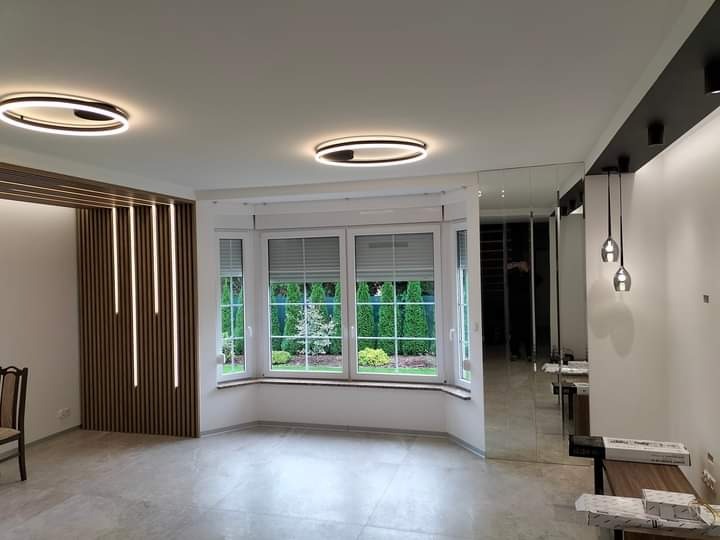 Nowoczesne wnętrze po remoncie: okno wykuszowe z żaluzjami, dekoracyjna ściana z lameli z oświetleniem LED, lustra na ścianie i designerskie lampy sufitowe.