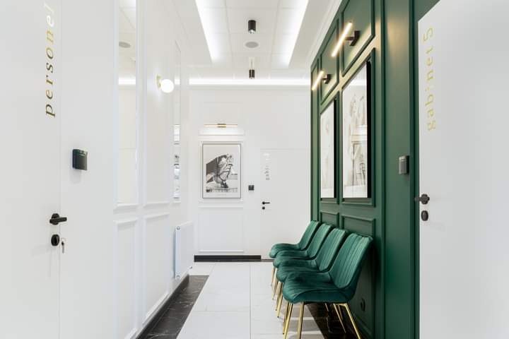 Korytarz w nowoczesnym stylu z zielonymi panelami ściennymi, białymi drzwiami z numerami gabinetów i eleganckimi, zielonymi krzesłami na złotych nogach.