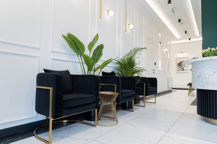 Eleganckie wnętrze poczekalni z czarnymi fotelami na złotych nogach, marmurową podłogą i ścianami z białymi panelami. Rośliny doniczkowe dodają świeżości.