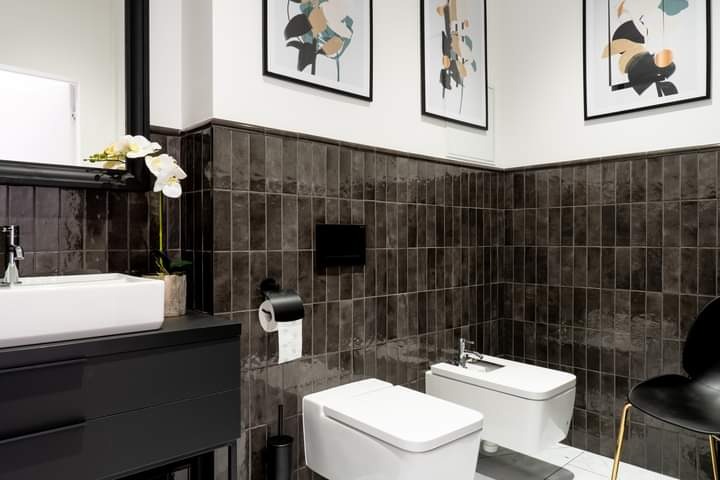 Nowoczesna łazienka z ciemnymi, błyszczącymi płytkami ułożonymi pionowo do połowy ściany, białą armaturą i czarnymi akcentami, takimi jak rama lustra i krzesło.