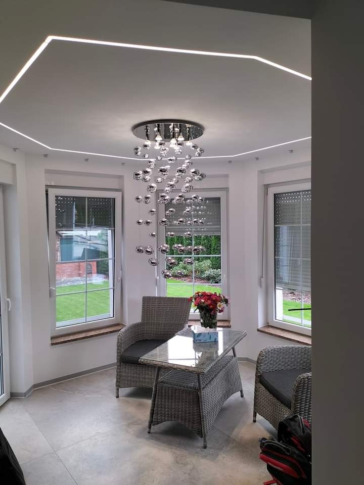 Jasne wnętrze z oświetleniem LED na suficie, szklanym stołem i wiklinowymi krzesłami. Widoczne okna z widokiem na zieleń. Nowoczesny design i wykończenie.