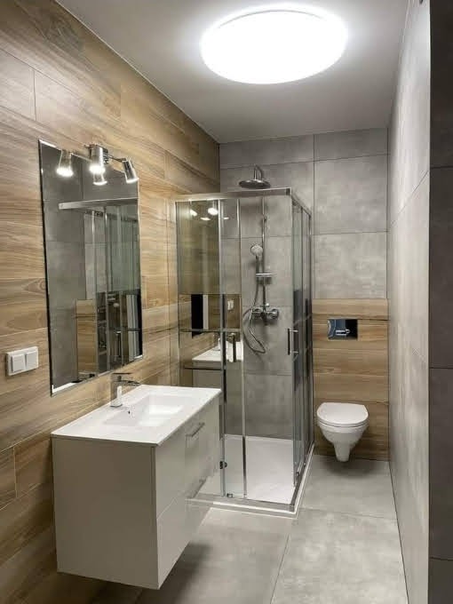 Nowoczesna łazienka z kabiną prysznicową, umywalką na szafce i lustrem. Ściany wyłożone płytkami imitującymi drewno i szarym gresem. Oświetlenie sufitowe.